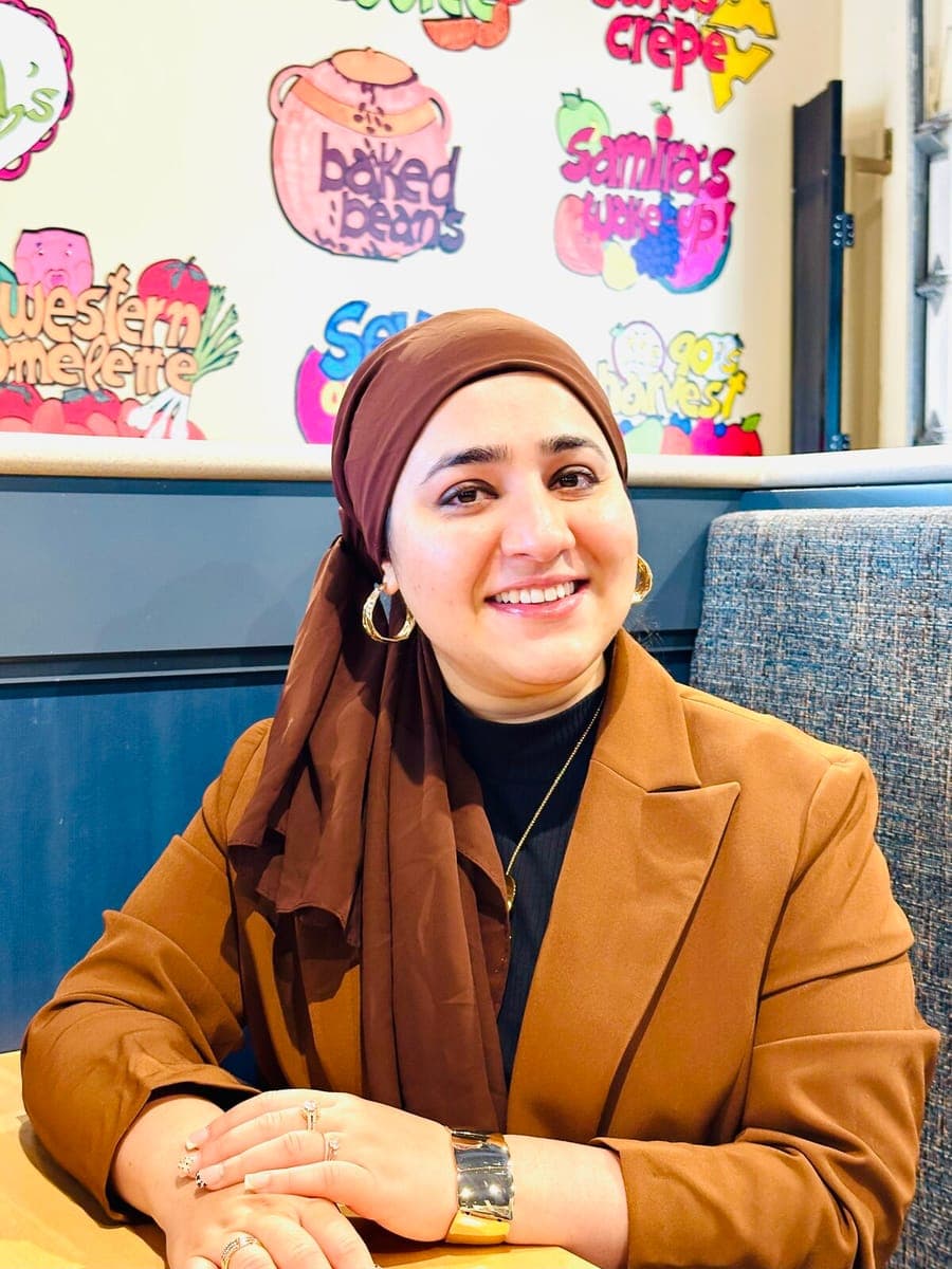Seemab Akbar at a café table, smiling, in a brown blazer and headscarf