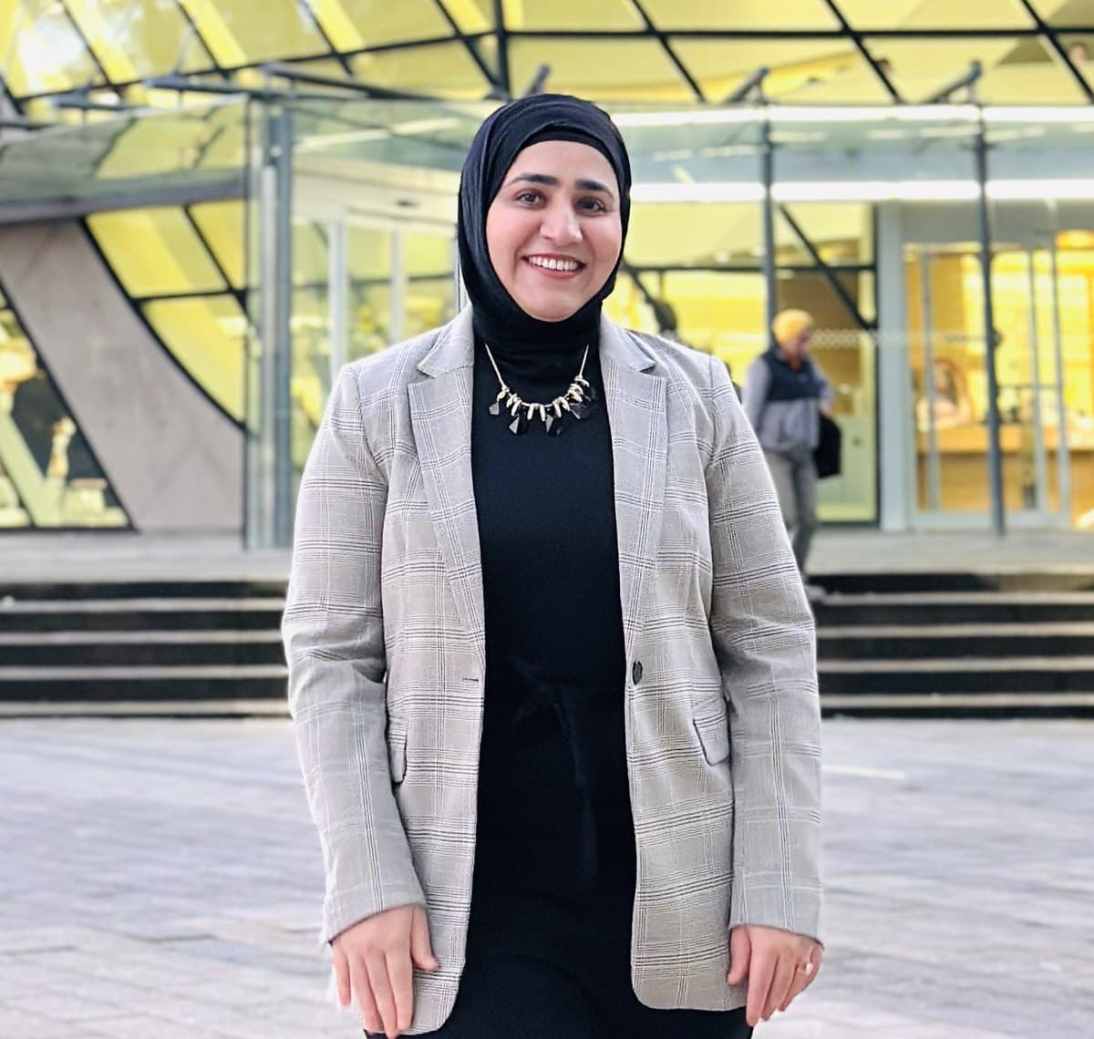 Seemab Akbar smiling on a city plaza in the evening, in a grey check blazer and black hijab